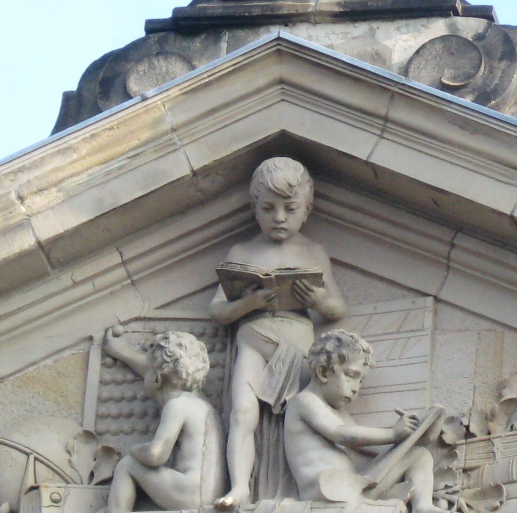 Giebel der Pestalozzisschule mit plastischen Figuren: Es sind Schulkinder zu sehen, die von links nachrechts – rechnen lesen schreiben.